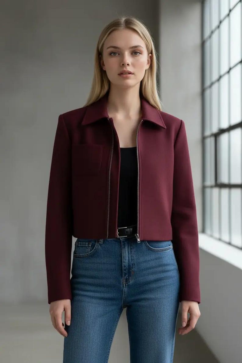 May include: A burgundy cropped jacket with a collared neckline and a silver zipper. The jacket has a single pocket and is paired with blue jeans and a black top. The model is standing in front of a window.