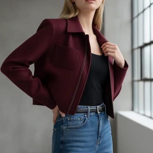 May include: A burgundy cropped jacket with a collared neckline and a front zipper. The jacket has a single pocket on the left side and is paired with a black top and blue jeans. A black belt is worn at the waist.