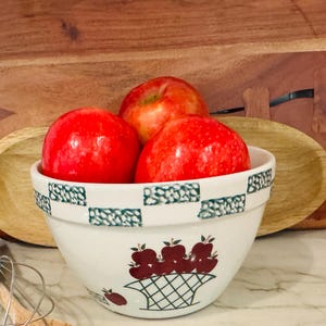 May include: A white ceramic bowl with a green and white geometric pattern and a basket of apples design, filled with three red apples. The bowl is on a marble surface, with wooden cutting boards in the background.