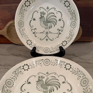 May include: Two white ceramic plates with a green rooster design in the center. The plates have a decorative border with floral and leaf patterns. The plates are displayed on a black stand.