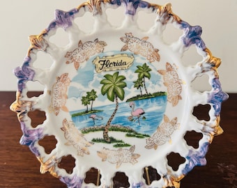 May include: A decorative porcelain plate with a lattice-style scalloped edge featuring purple and gold trim. The center depicts a Florida scene with palm trees, pink flamingos, and blue water, surrounded by a border of gold-outlined orange fruit.