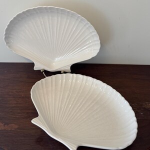 May include: Two white ceramic dishes shaped like scallop shells. Each dish features a glossy finish and a series of raised, radiating ridges that extend from the hinge area to the scalloped outer edge, creating a textured, fan-like appearance.