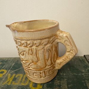 May include: A tan ceramic pitcher with a decorative embossed design. The pitcher has a spout and a handle with a hole. The interior is a lighter tan color. The pitcher is sitting on a green surface.