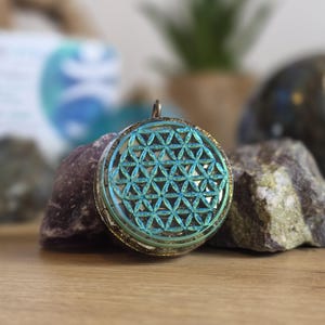 May include: A round pendant with a teal and gold design featuring a geometric pattern. The pendant is set against a backdrop of rocks and a wooden surface, with a blurred background.