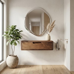 Puede incluir: Una consola de caj&oacute;n flotante montada en la pared con una forma rectangular redondeada y lisa con un acabado de grano de madera natural. Sobre la consola cuelga un espejo de forma org&aacute;nica irregular con un marco de plata pulida que refleja.