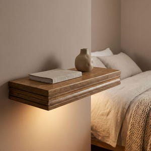 Puede incluir: Una estantería flotante de madera con un acabado cálido y natural. Un pequeño jarrón beige y un libro descansan sobre la estantería. La estantería está montada en una pared de color neutro, con una cama al fondo. La estantería está iluminada desde abajo.