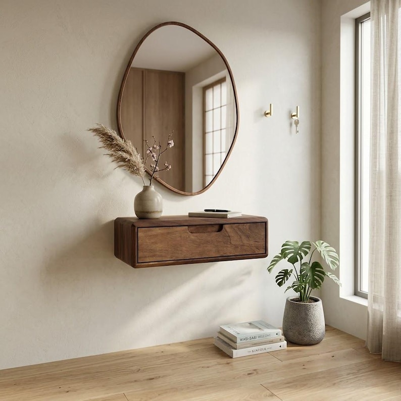 Puede incluir: Una consola flotante de madera oscura montada en la pared con un caj&oacute;n de tirador horizontal. Encima cuelga un espejo de forma org&aacute;nica irregular con un marco fino de madera oscura. Un peque&ntilde;o jarr&oacute;n beige con follaje seco descansa sobre la consola.