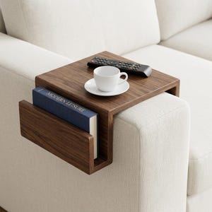 Op de afbeelding: Een donkerbruine houten sofa-armtafel met een ingebouwde boekenhouder. Een witte kop en schotel staan op de tafel, samen met een afstandsbediening. Een blauw boek met de tekst "MODERN LEISURE" staat in de houder.