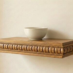 May include: A light-colored ceramic bowl sits on a rectangular, wooden shelf. The shelf is a warm brown color and features a decorative, carved design along the front edge. The shelf is mounted on a neutral-colored wall.
