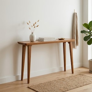 May include: A rectangular wooden console table with four tapered legs. On the surface sits a white ceramic vase containing dried stems and two stacked books. The table features a smooth, light brown wood grain finish and a clean, simple design.