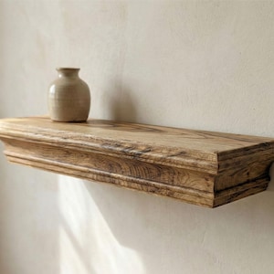 Puede incluir: Estante flotante de madera rústica con borde decorativo, montado en una pared de color neutro. Un pequeño jarrón de cerámica beige está encima. El estante mide aproximadamente 76 cm de largo.