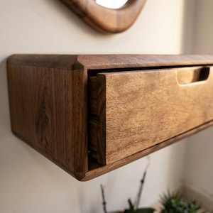 Puede incluir: Un estante flotante montado en la pared hecho de madera de color marr&oacute;n oscuro con un patr&oacute;n de grano visible. El estante cuenta con un caj&oacute;n rectangular &uacute;nico con un tirador empotrado integrado en el borde superior y esquinas redondeadas.