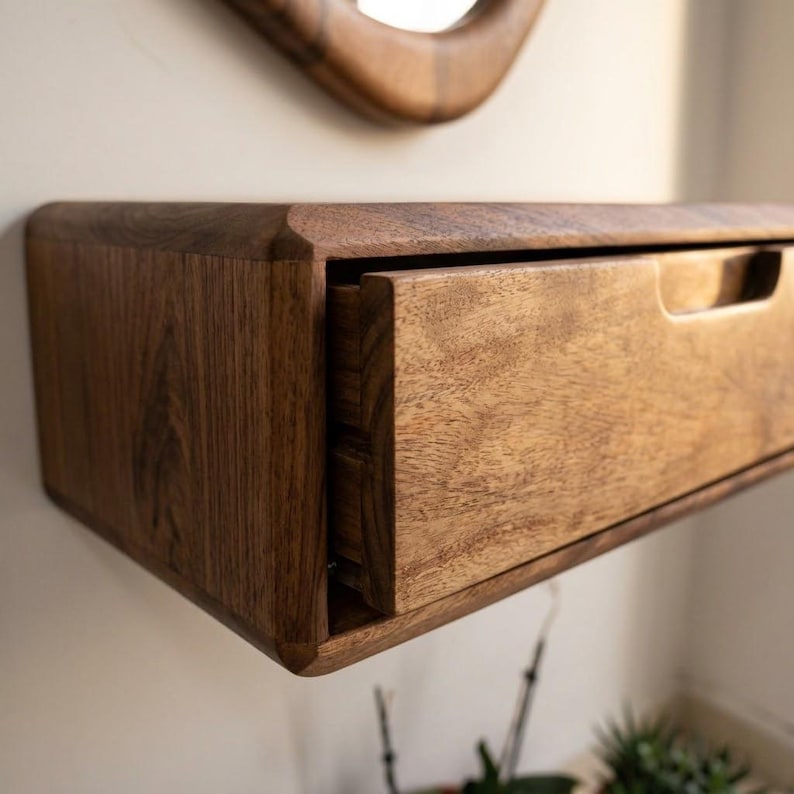Puede incluir: Un estante flotante montado en la pared hecho de madera de color marr&oacute;n oscuro con un patr&oacute;n de grano visible. El estante cuenta con un caj&oacute;n rectangular &uacute;nico con un tirador empotrado integrado en el borde superior y esquinas redondeadas.