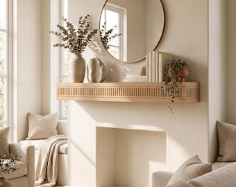 May include: A light wood fireplace mantel with a vertical slat design, mounted on a cream wall. Above the mantel sits a round gold-framed mirror, a ceramic vase with eucalyptus branches, a stone sculpture, books, and a trailing green plant.