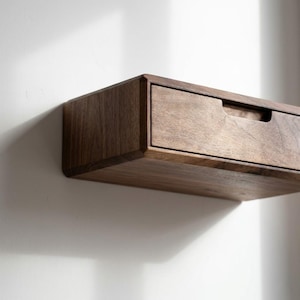 Puede incluir: Un caj&oacute;n flotante montado en la pared hecho de madera marr&oacute;n oscura con una veta natural visible. La unidad rectangular cuenta con un solo caj&oacute;n frontal con un recorte de asa empotrado a lo largo del borde superior, montado contra una pared blanca.