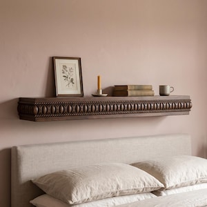 Puede incluir: Un estante flotante de madera marrón oscuro con un patrón tallado en forma de cuerda a lo largo del borde frontal. El estante está montado en una pared y sostiene una lámina botánica enmarcada, una vela amarilla, dos libros y una taza.
