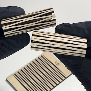 May include: Close-up of three wooden objects with a light brown color and dark brown etched lines. Two are cylindrical, and one is rectangular with the number "1.5" etched on it. The objects are held by a person wearing black gloves.
