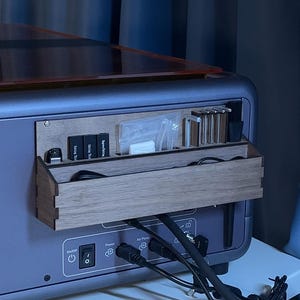 Puede incluir: Una impresora 3D gris con un organizador de madera adjunto. El organizador contiene varias herramientas, incluyendo botellas pequeñas, pinceles y recipientes etiquetados. Los puertos de alimentación y bomba de aire son visibles en la parte trasera de la impresora.