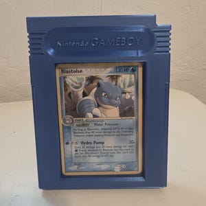 May include: A blue 3D-printed Game Boy cartridge case holding a Pokémon trading card. The card features Blastoise with 120 HP and the text "Hydro Pump". The case is labeled "Nintendo GAMEBOY".