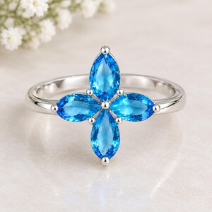 Pode incluir: Um anel de prata com quatro pedras preciosas azul-brilhante em corte marquise, dispostas num padrão de cruz ou flor. A banda é de prata polida e as pedras são mantidas no lugar por pequenas garras de metal, criando um design simétrico.