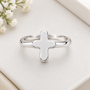 Puede incluir: Un anillo de tono plateado pulido que presenta una pieza central en forma de cruz con bordes suaves y redondeados. La banda es delgada y circular, conectándose al elemento central de la cruz que tiene una superficie metálica reflectante.