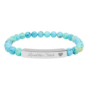 Op de afbeelding: Een kralenarmband met turquoise en lichtgroene kralen. Een zilverkleurige balk in het midden is gegraveerd met de woorden "Loved like Stitch" en een hartsymbool. De armband is ontworpen om om de pols te worden gedragen.