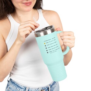 May include: A light blue insulated tumbler with a handle and a silver lid. The tumbler has a straw and features text that reads "Progress Report 2020-2025" and other phrases. The tumbler is held by a person.