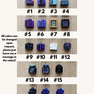 May include: A collection of eighteen custom mechanical keyboard keycaps arranged in rows, featuring designs such as miniature books, dragons, picture frames, candles, potion bottles, birdcages, and crystals in various colors including purple, blue, and black.