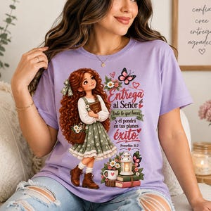 May include: A light purple t-shirt featuring a graphic of a girl with long curly red hair wearing a green floral dress and brown boots. The design includes a lantern, books, flowers, butterflies, and the Spanish text: Entrega al Señor todo lo que haces y él pondrá en tus planes éxito. Proverbios 16:3.