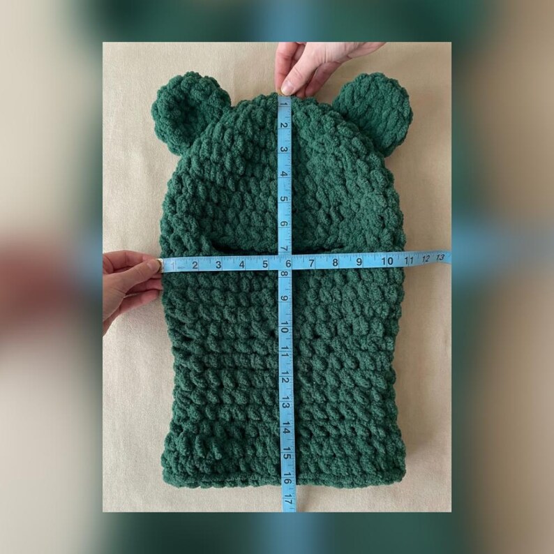 May include: A dark green, handmade balaclava-style hat with bear ears. The hat is measured with a blue measuring tape, showing a width of approximately 6 inches and a height of approximately 17 inches.