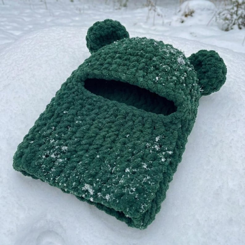 May include: A dark green, knitted balaclava with ear-shaped embellishments. The face opening is visible, and the hat is covered in small snowflakes. The balaclava is resting on a snowy surface.