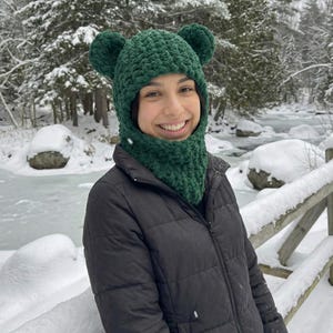 May include: A green crocheted balaclava with bear ears. The balaclava covers the head, neck, and part of the face. The person in the image is wearing a black jacket and is smiling. The background shows a snowy landscape with trees and a river.