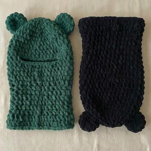 May include: Two handmade, knitted balaclavas. One is forest green with bear ears and a face opening. The other is navy blue with a textured knit and a rounded bottom. Both are made of a soft, chunky yarn.