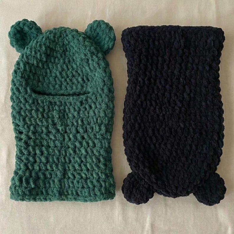 May include: Two handmade, knitted balaclavas. One is forest green with bear ears and a face opening. The other is navy blue with a textured knit and a rounded bottom. Both are made of a soft, chunky yarn.