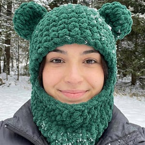May include: A green crocheted balaclava with bear ears. The balaclava covers the head, neck, and lower face, with ear-shaped protrusions on top. The textured yarn is a vibrant green color, and the background shows a snowy outdoor scene.