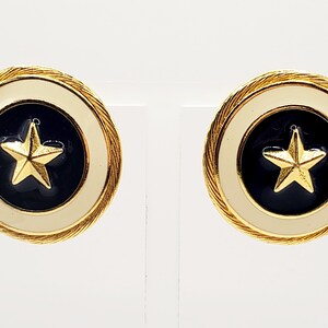Puede incluir: Par de pendientes de estrella dorados. Cada pendiente presenta una estrella dorada sobre un fondo azul marino, rodeada por un borde crema y un borde trenzado dorado. Sobre un fondo blanco.