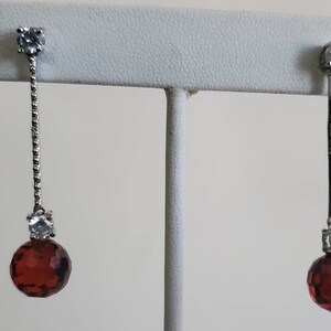 Op de afbeelding: Hangende oorbellen met een zilverkleurig metalen ontwerp. Elke oorbel heeft een heldere, vierkant geslepen steen aan de bovenkant en een gefacetteerde, ronde, donkerrode steen aan de onderkant. De oorbellen worden op een witte standaard getoond.