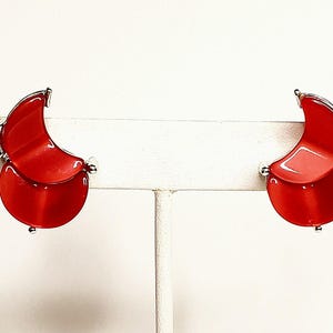 May include: A pair of red crescent moon shaped earrings with silver accents. The earrings are on a white display stand.