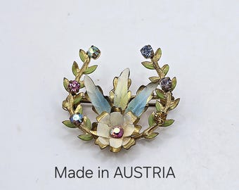 May include: A gold-toned brooch with a white flower and blue and green leaves. The brooch is decorated with rhinestones and has the text "Made in AUSTRIA" on the bottom.