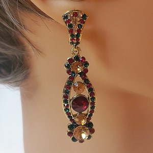 Pode incluir: Brincos de argola dourados com pedras de imitação vermelhas, verdes e transparentes. Os brincos têm a forma de uma lágrima com um cacho de pedras de imitação na parte inferior.
