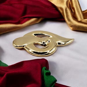 Può includere: Un fermaglio in metallo dorato a forma di nodo o vortice stilizzato. Il fermaglio ha una finitura lucida e lucidata.