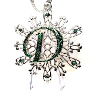 May include: A silver snowflake ornament with a green glitter letter D in the center. The ornament is hanging from a silver chain and has a clear plastic stand.