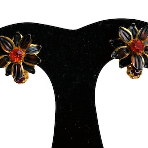 May include: A pair of gold tone earrings with black flower-shaped designs and red and orange rhinestones.