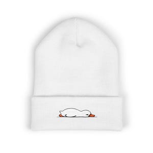 Puede incluir: Gorro blanco con un diseño de pato de dibujos animados bordado. El pato está representado tumbado, con pies y pico naranjas. Este gorro informal es un accesorio versátil para el uso diario.