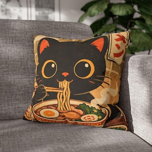 Op de afbeelding: Vierkant kussen met een zwarte kat die ramen eet. De kat heeft grote gele ogen en oranje oren. De ramenkom bevat noedels, een ei en groenten. Het kussen heeft een vintage, geïllustreerde stijl.