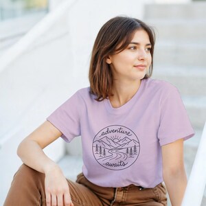 Könnte beinhalten: Ein helllila T-Shirt mit einem schwarzen, kreisförmigen Aufdruck mit der Aufschrift "adventure awaits". Das Design zeigt Berge, eine Sonne, Bäume und einen Fluss. Das T-Shirt ist aus weichem Material.