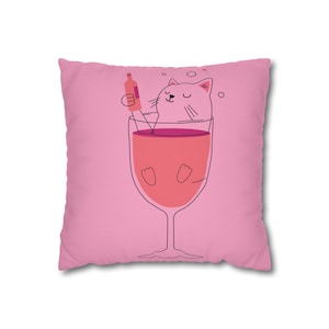 May include: Pink square pillow with a cartoon illustration of a cat relaxing in a wine glass. The cat is pink with black whiskers and is holding a wine bottle. The wine glass is filled with pink liquid.