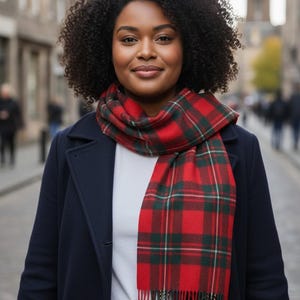 Puede incluir: Bufanda a cuadros rojos y verdes con flecos, sobre un abrigo azul marino. El patrón de cuadros es un tartán clásico. La imagen está tomada al aire libre, con una calle de la ciudad borrosa en el fondo.