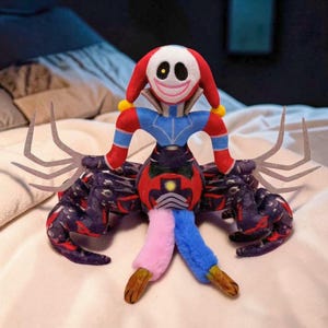 May include: A plush toy featuring a clown-like character with a white face, red hair, and a wide smile. The toy has a blue and red torso, a patterned body, and multi-colored legs. The toy has multiple gray, claw-like appendages.