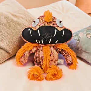 May include: A plush monster toy with a patterned body in shades of orange, pink, and brown. It has orange fur accents, large white teeth, and round, blue-gray eyes. The toy is sitting on a white surface.